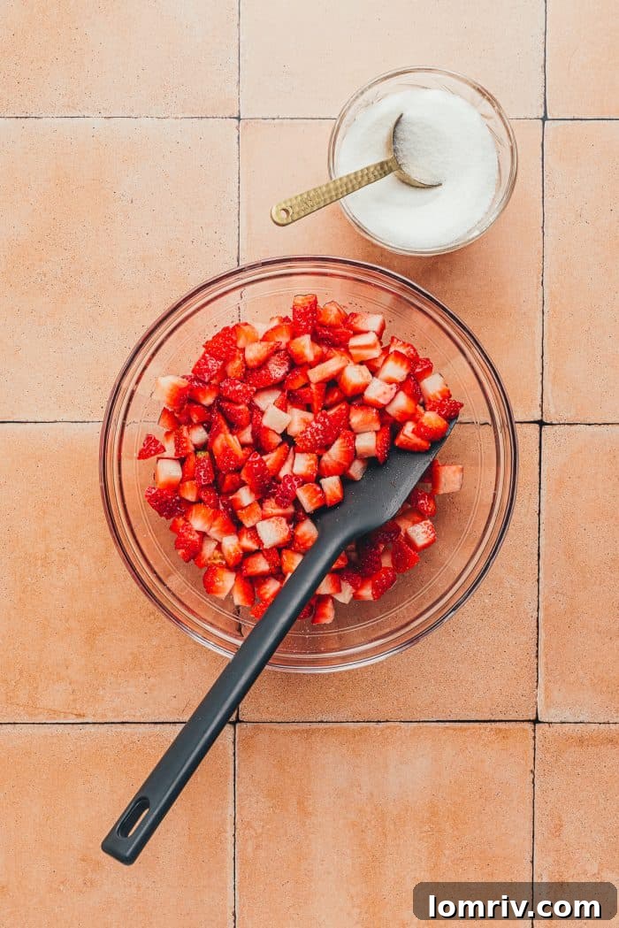 A glass bowl with diced strawberries and a black spatula. 