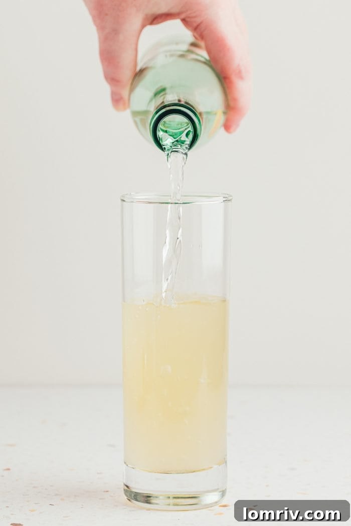 Adding sparkling mineral water to the elderflower and citrus mixture in a tall glass.