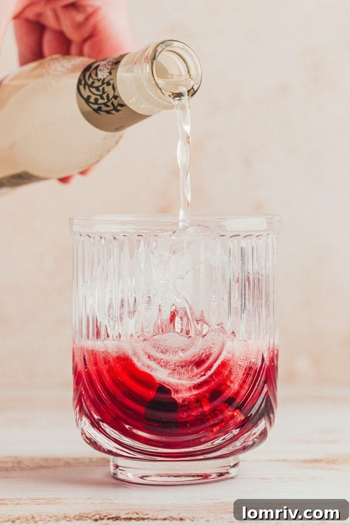 A glass with muddled cherries, lime juice, and ginger beer being poured into it.