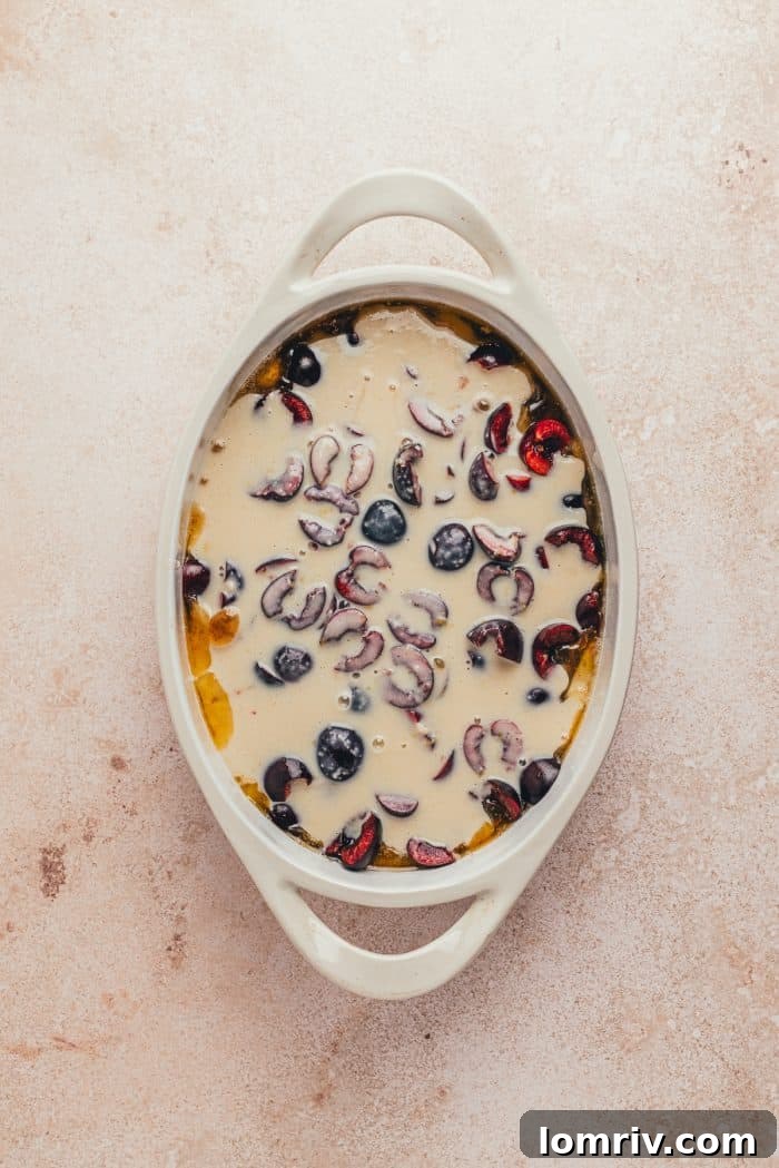 A white oval dish with sliced cherries in it and cobbler batter poured over it.