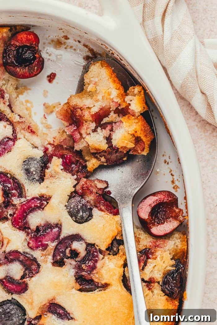 An oval baking dish with cherry cobbler and a spoon with a scoop of cobbler.