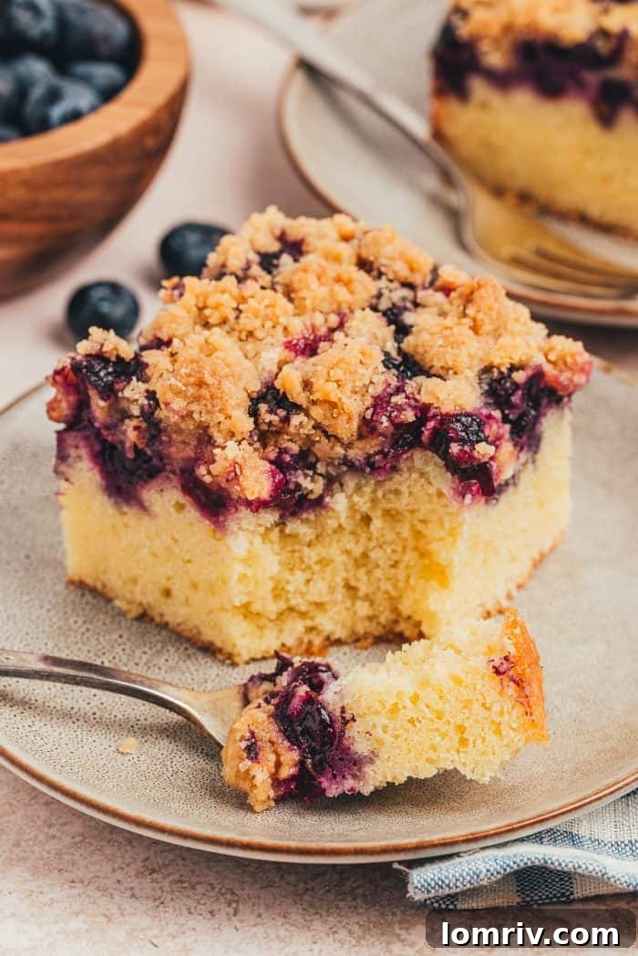 A close-up of a blueberry crumb cake slice on a small plate, with a fork digging in for a bite, emphasizing its delicious layers.