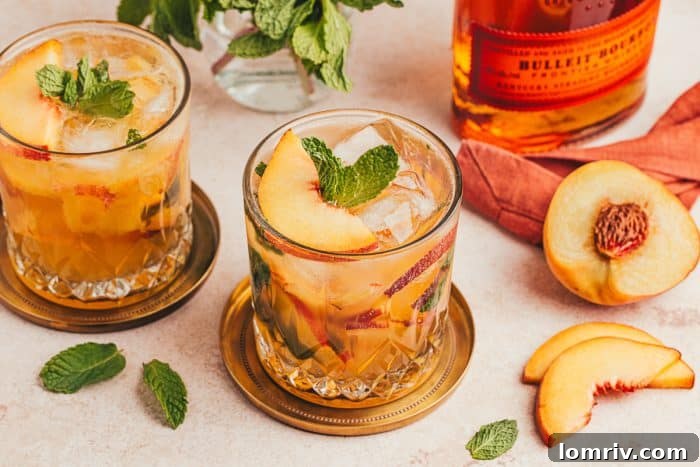 Two glasses filled with a bourbon peach smashes with peaches in the background and a bottle of bourbon.