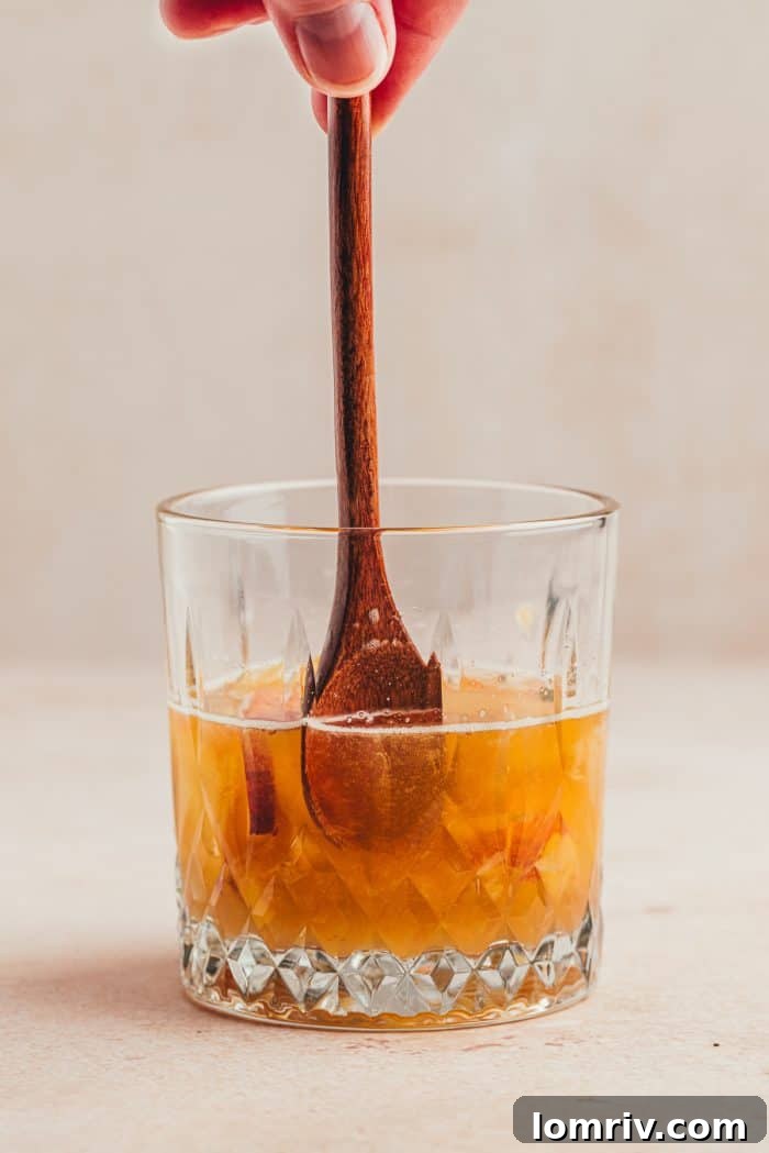 A glass with a wooden spoon and the ingredients for a peach bourbon smash.
