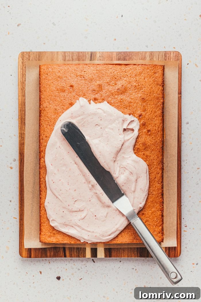 A baked cake being generously frosted with creamy strawberry frosting, creating a smooth, even layer