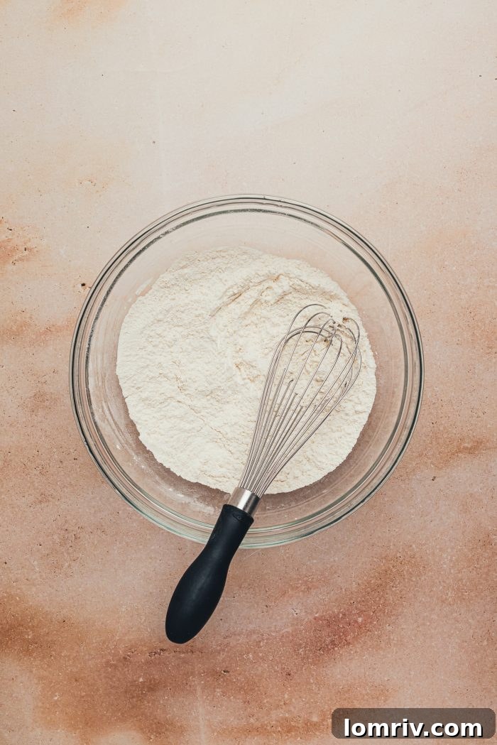 A glass bowl containing dry ingredients, including flour, with a whisk resting inside.