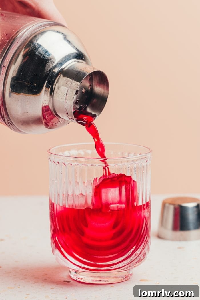 A cocktail shaker pouring the vibrant blueberry gin mixture into a double rocks glass filled with fresh ice.