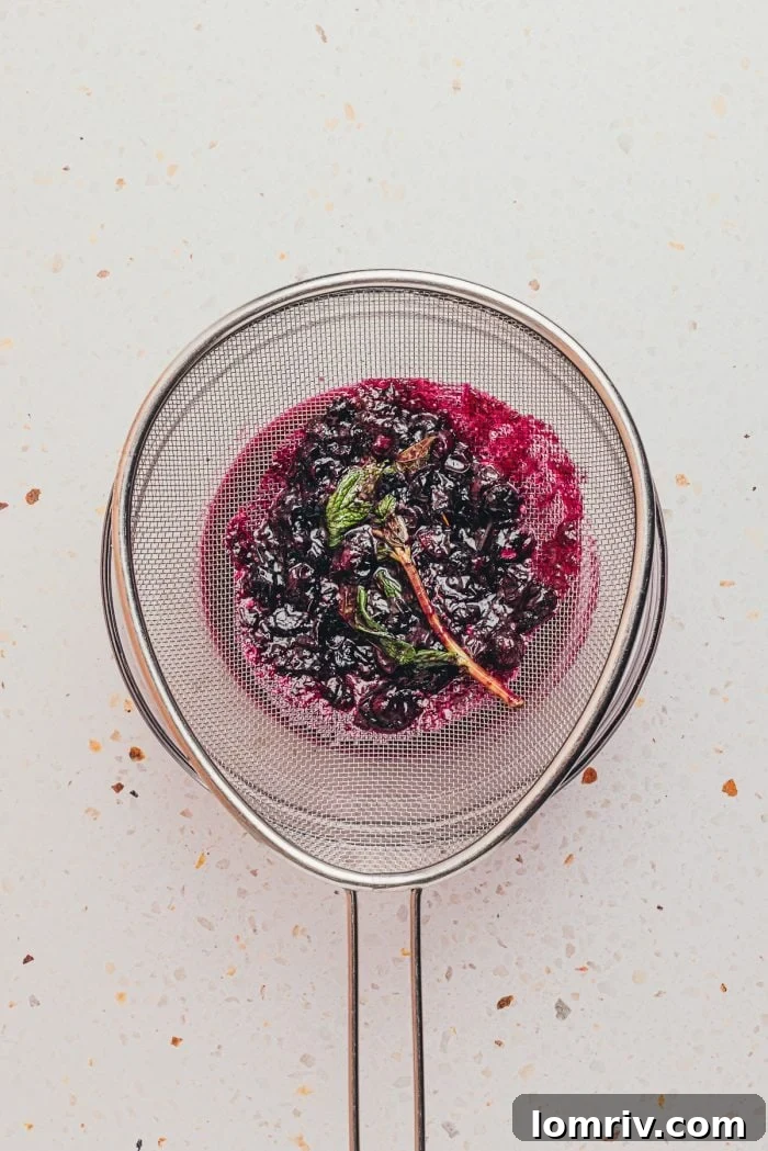 A fine-mesh sieve straining homemade blueberry simple syrup into a glass container.