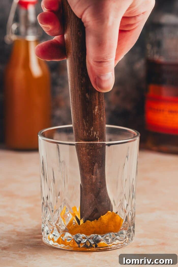 A muddling stick actively pressing the orange peel, bitters, and syrup together in a glass, releasing aromas.