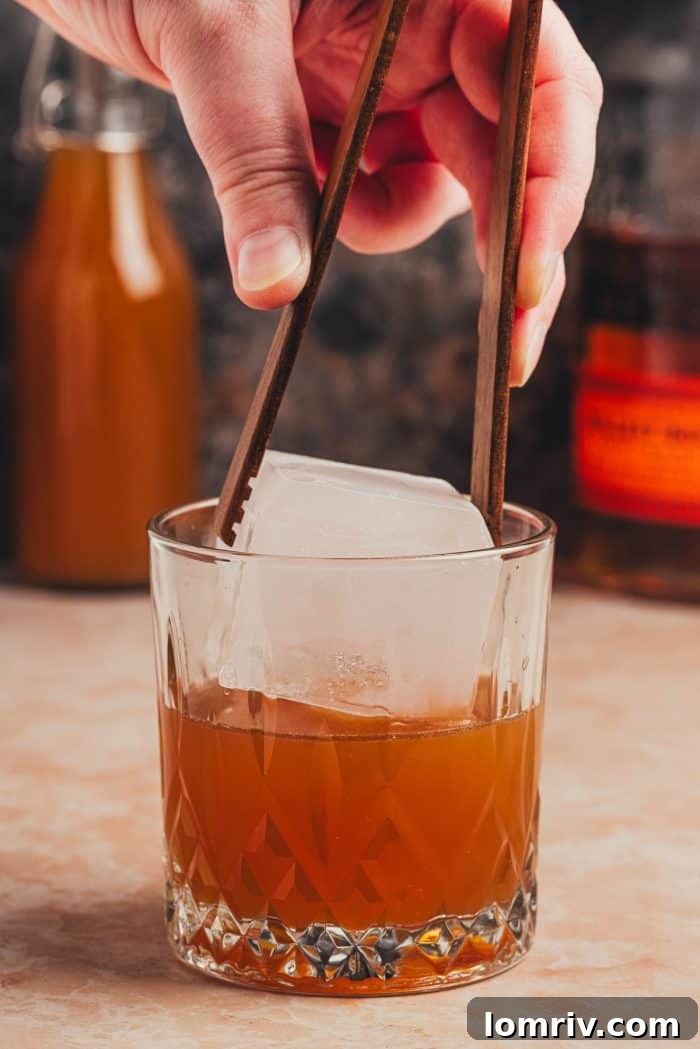 A large ice cube being carefully placed into the Apple Cider Old Fashioned glass, signaling the final step before serving.