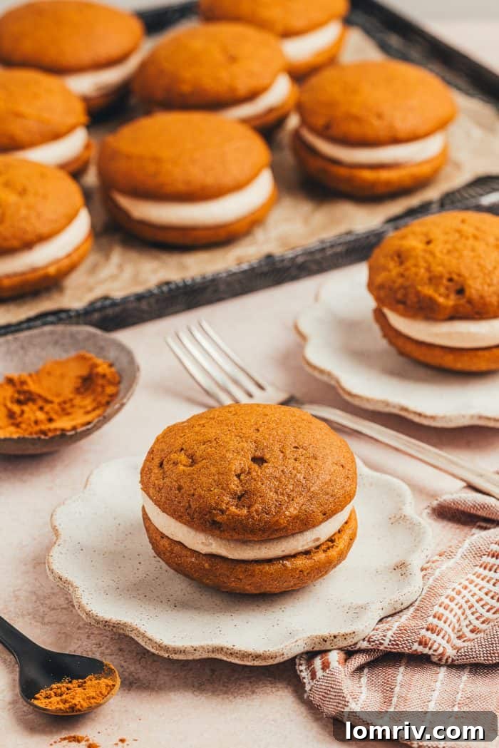 Delightful pumpkin whoopie pies with cinnamon cream cheese filling on a ruffled white plate, with a baking tray full of more pies in the background.