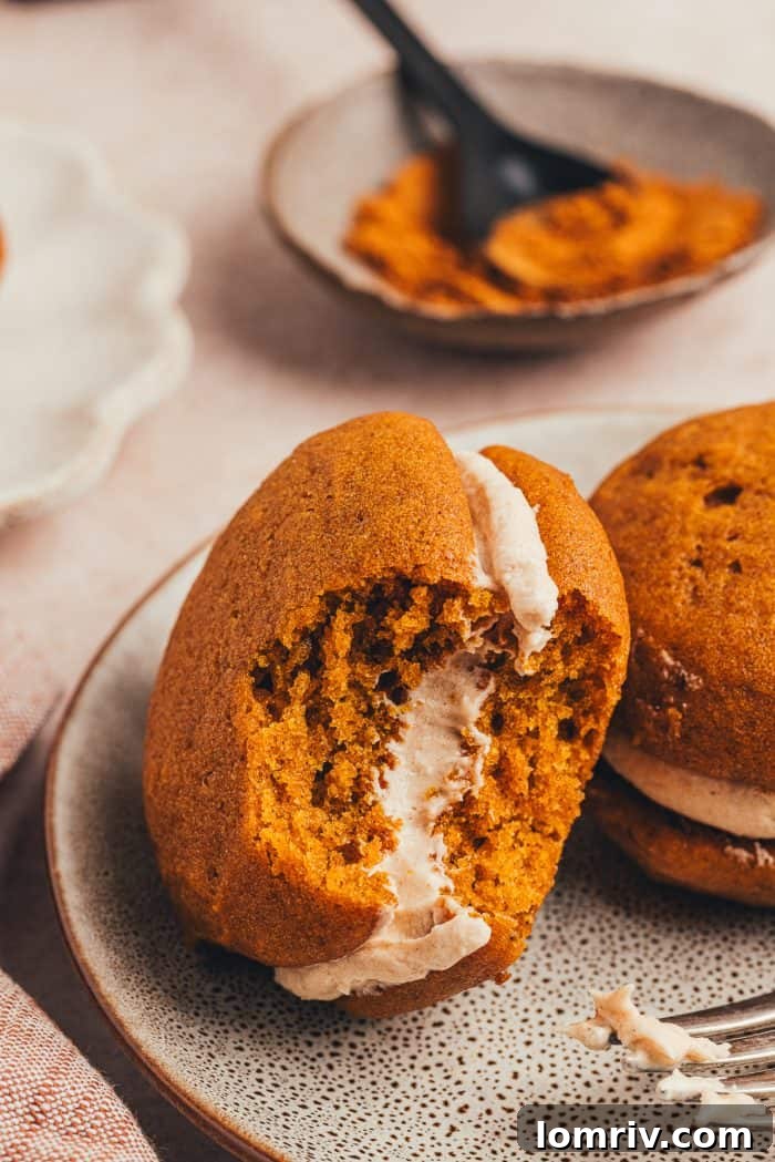 A pumpkin whoopie pie with a delicious bite taken out, resting on a plate next to another whole whoopie pie.