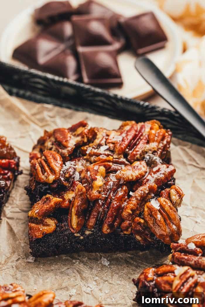 A single pecan pie brownie is on parchment paper with chocolate in the background.