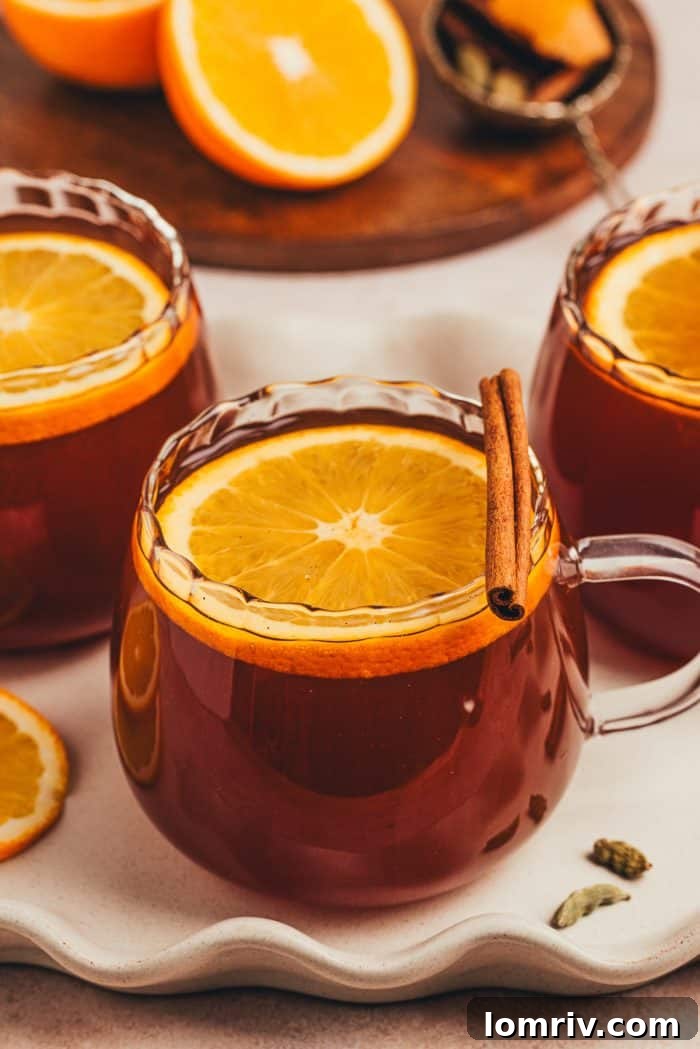 Two glass mugs, elegantly presented, filled with chai vanilla bean hot toddy and garnished with citrus, on a rustic surface.