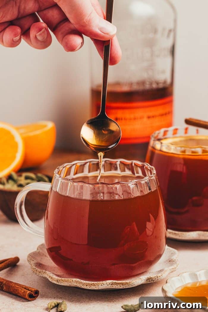 A spoon gently dipping into a glass mug of chai vanilla bean hot toddy, highlighting its inviting warmth and amber hue.