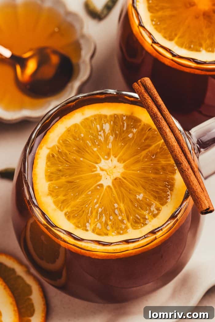 A close-up of a glass mug filled with chai vanilla bean hot toddy, garnished with a vivid orange slice, against a soft-focused background.