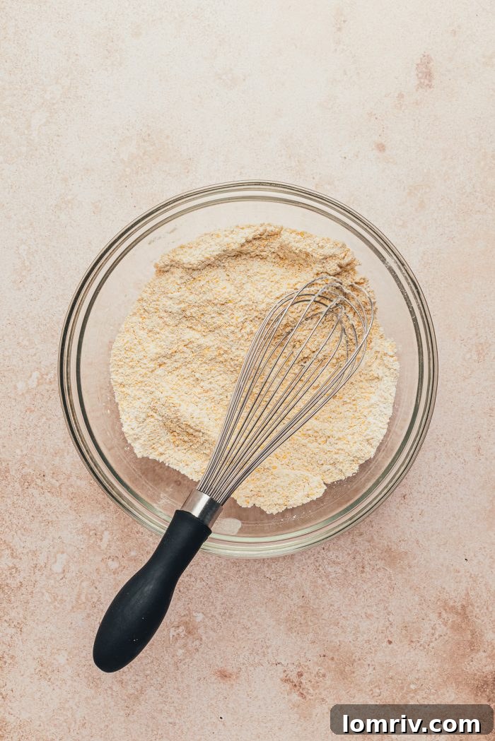 A glass bowl with cornmeal and flour and a whisk.