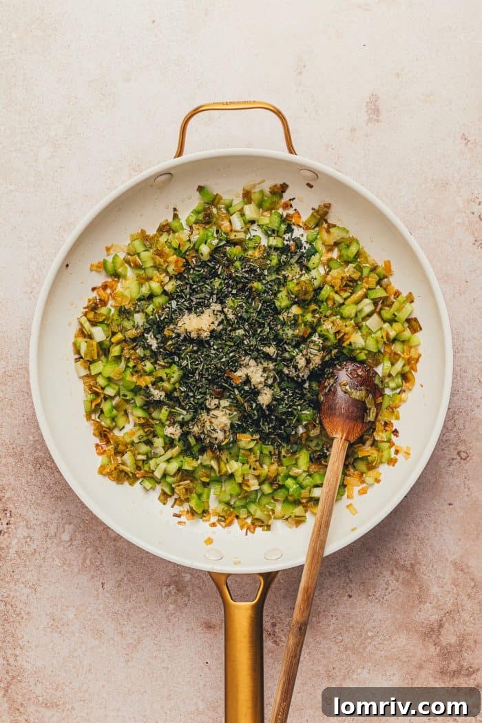 A white skillet with the leeks, celery, garlic, and herbs and a wooden spoon.