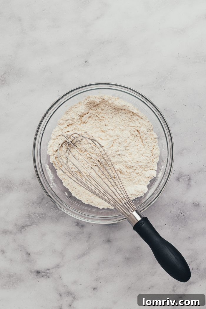 A glass mixing bowl with dry ingredients like flour and a whisk,