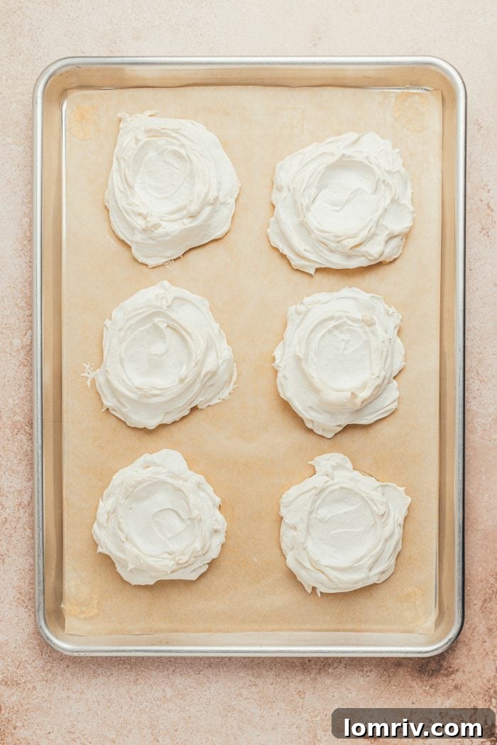 Baked mini pavlovas, golden and perfectly cracked, cooling on a baking sheet lined with parchment paper.