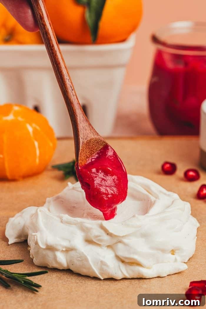 A spoon carefully ladling bright red cranberry curd into the hollow center of a freshly baked mini pavlova, creating a vibrant contrast.