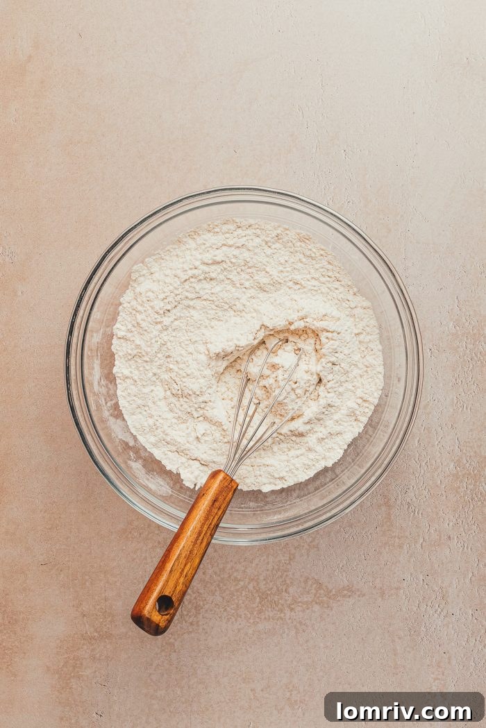 A glass bowl filled with the dry ingredients for the lemon pound cake, featuring a whisk resting inside, poised for mixing.