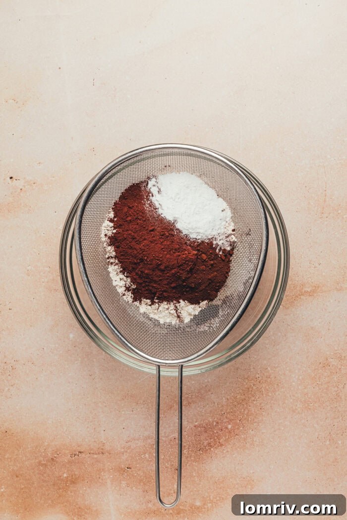 A close-up of dry ingredients, including flour, cocoa powder, and espresso powder, being carefully sifted into a large glass mixing bowl to ensure a smooth batter.