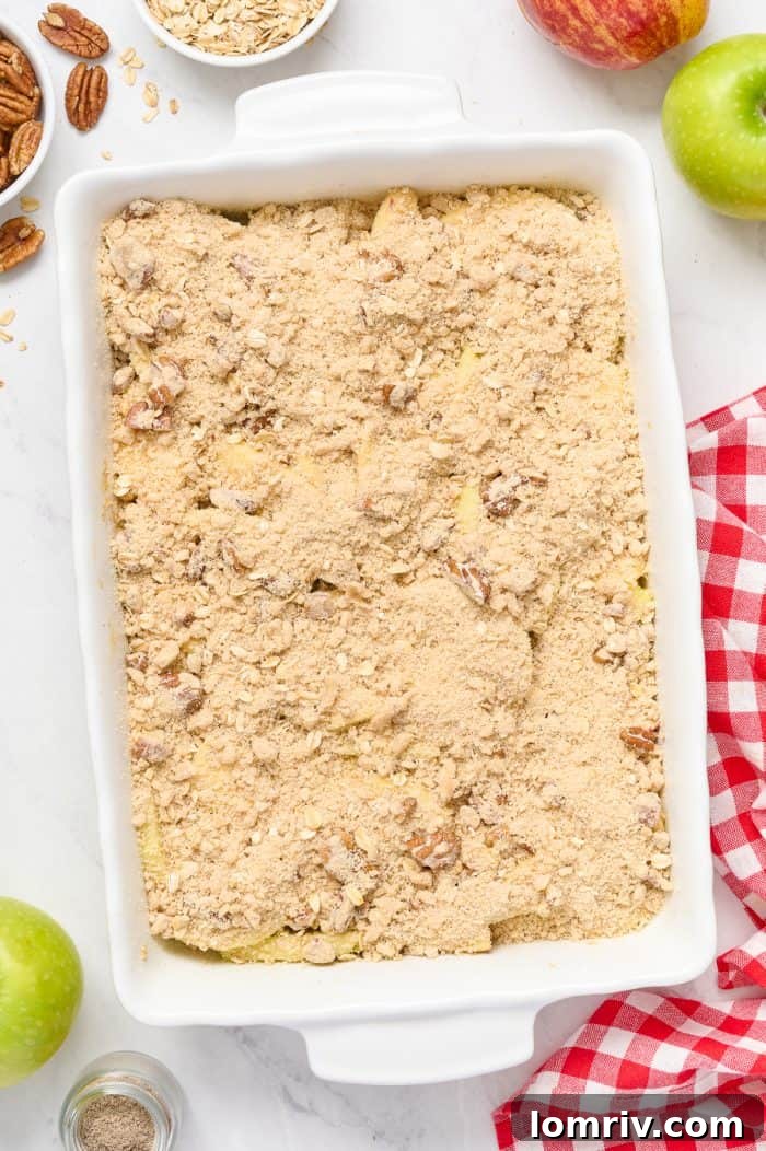 An unbaked apple cardamom crisp in a white baking dish, with the streusel topping generously scattered over the apples, poised for the oven.