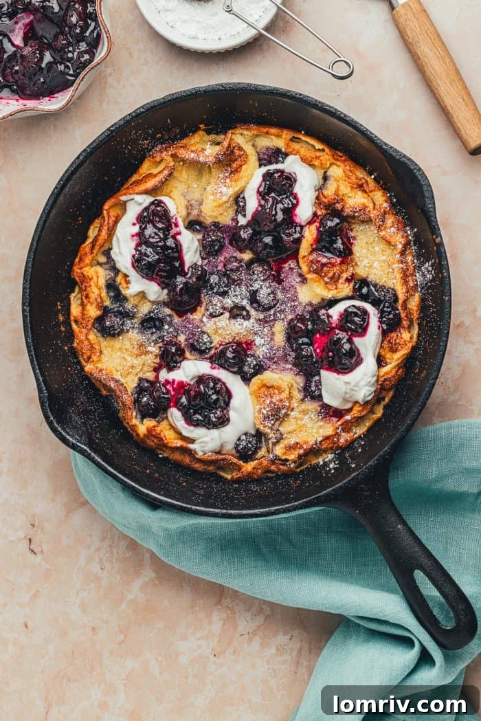 A skillet with a baked blueberry lemon Dutch baby pancake that is garnished with whipped cream and blueberry compote.