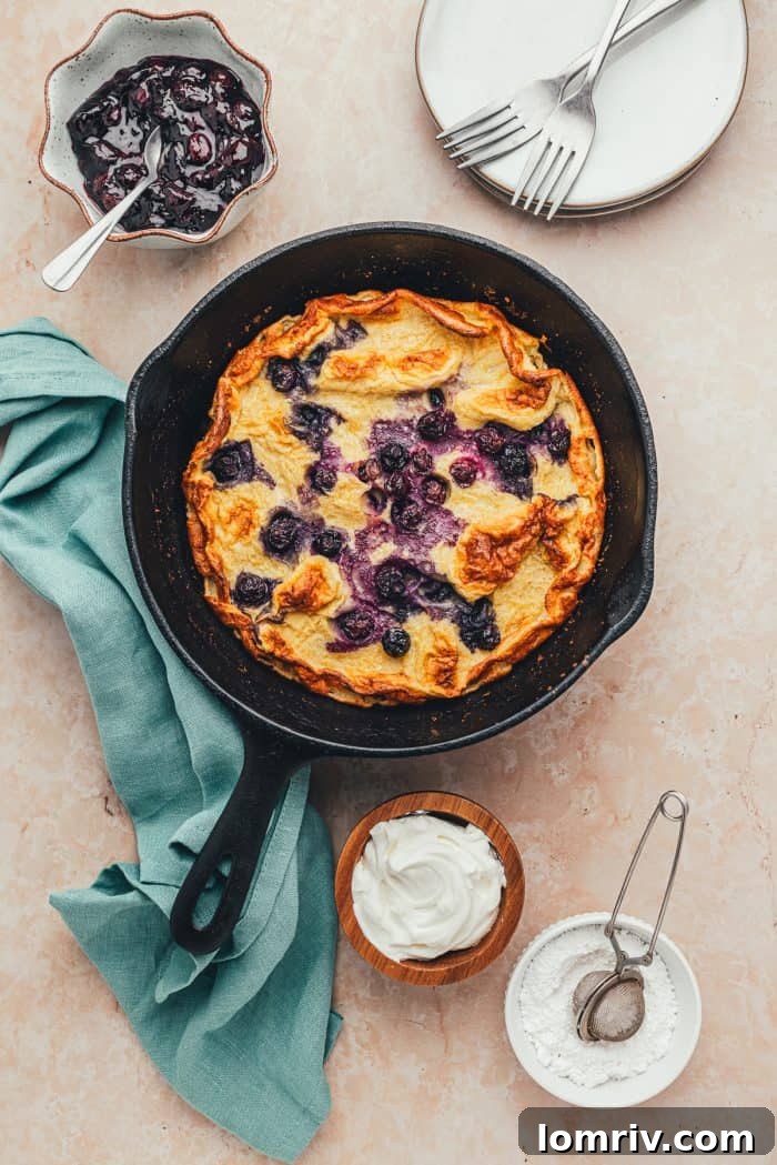 A skillet with a baked blueberry Dutch baby pancake. Bowls of garnishes and blueberry compote.