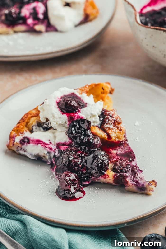 A plate with a slice of blueberry lemon Dutch baby pancake with a dollop of whipped cream and blueberry compote.