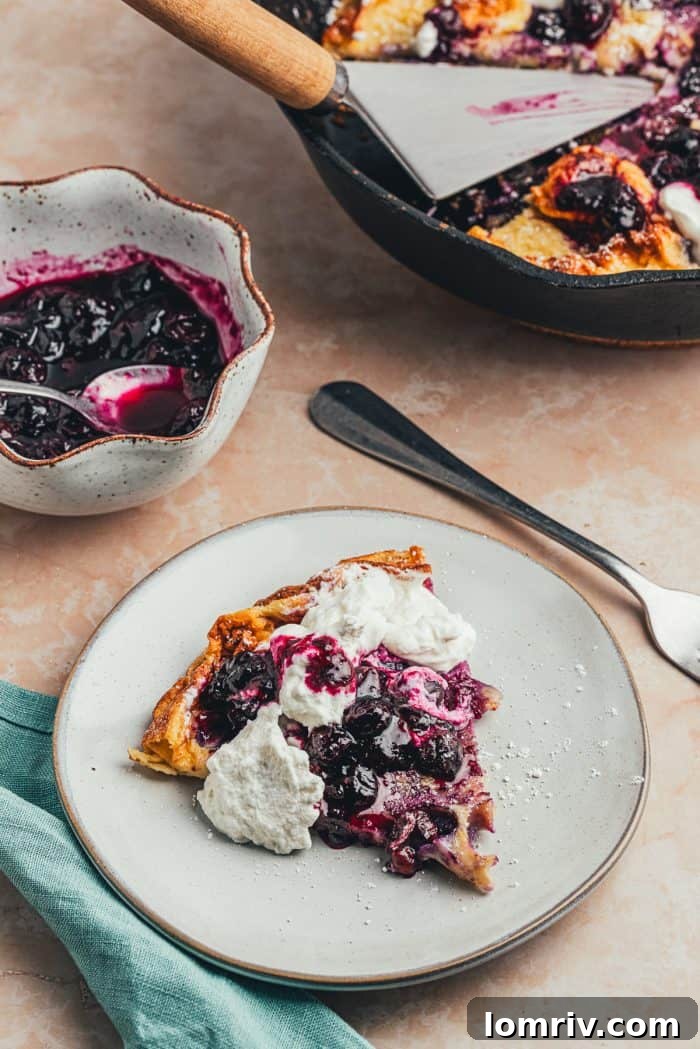 A plate with a slice of blueberry lemon Dutch baby pancake with a dollop of whipped cream and blueberry compote.