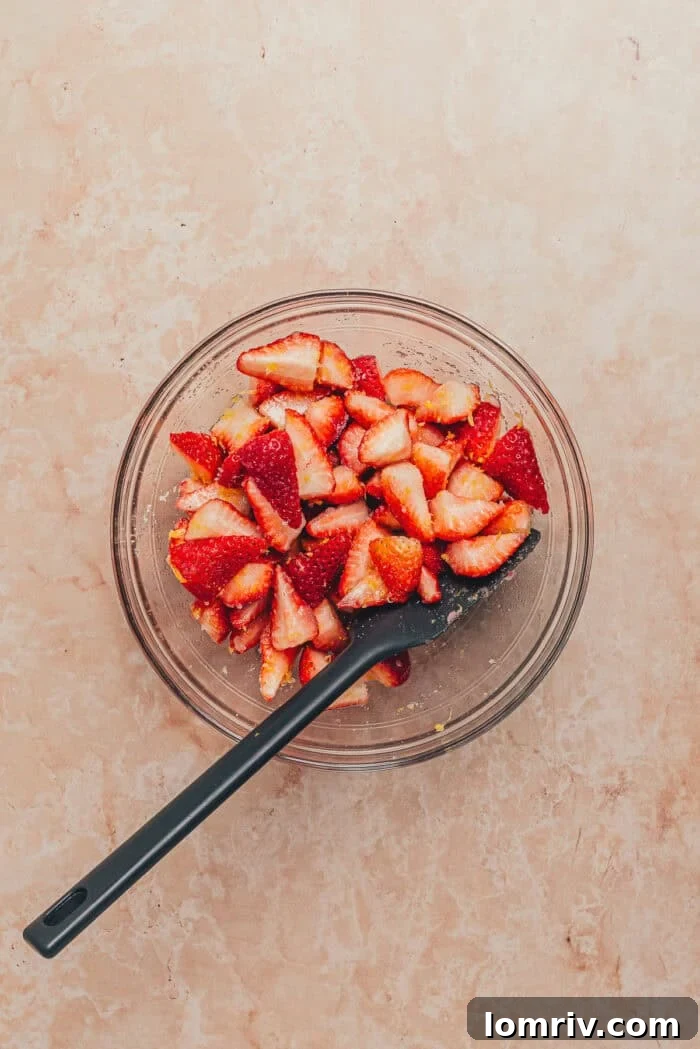 A bowl of freshly chopped strawberries being stirred with a spatula, with granulated sugar visible.