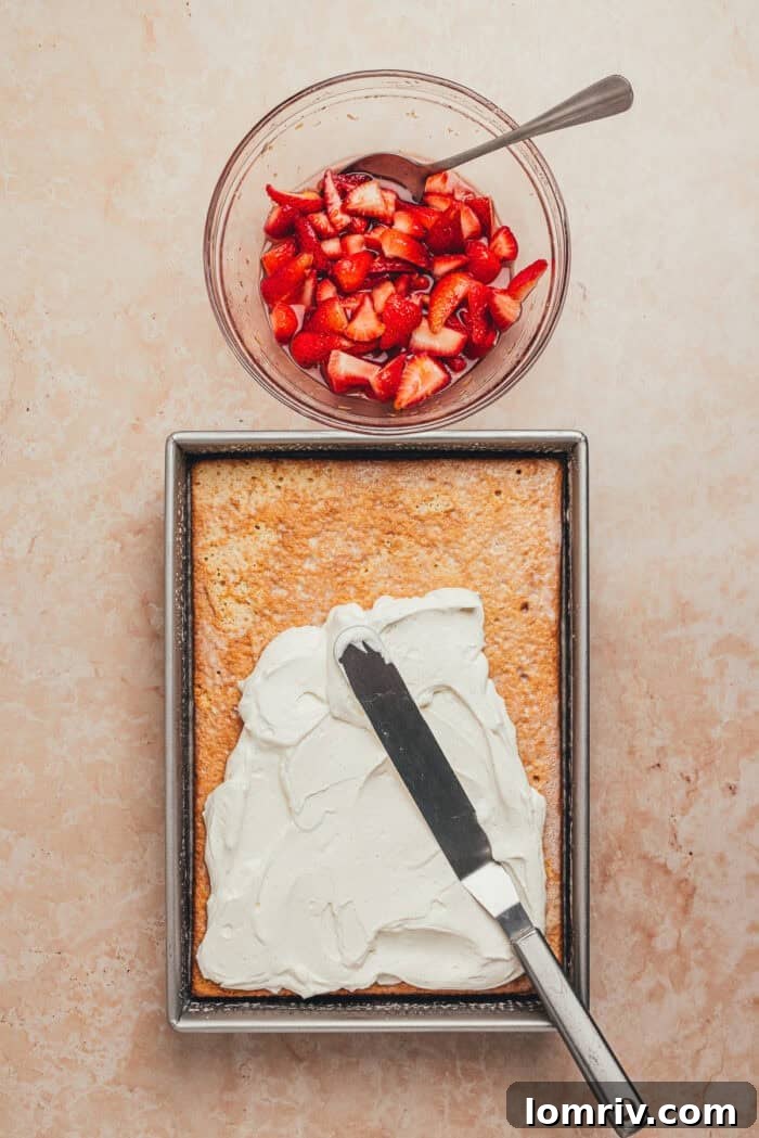 Whipped cream being artfully spread over the strawberry tres leches cake base in the pan, with a bowl of macerated strawberries nearby.
