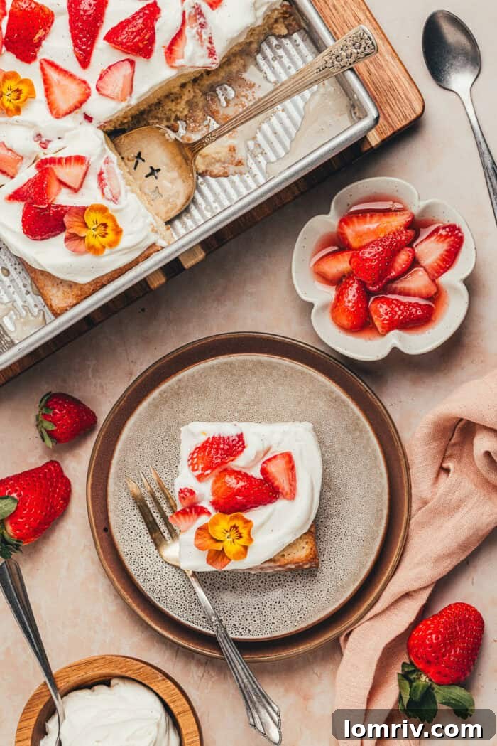 A slice of strawberry tres leches cake on a white plate, accompanied by a small bowl of fresh sliced strawberries.