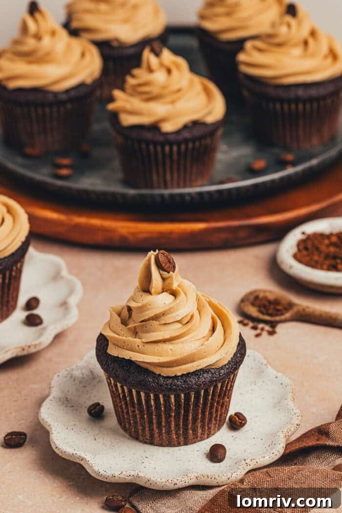 A single chocolate espresso cupcake on a white plate, with a platter of more cupcakes in the background.