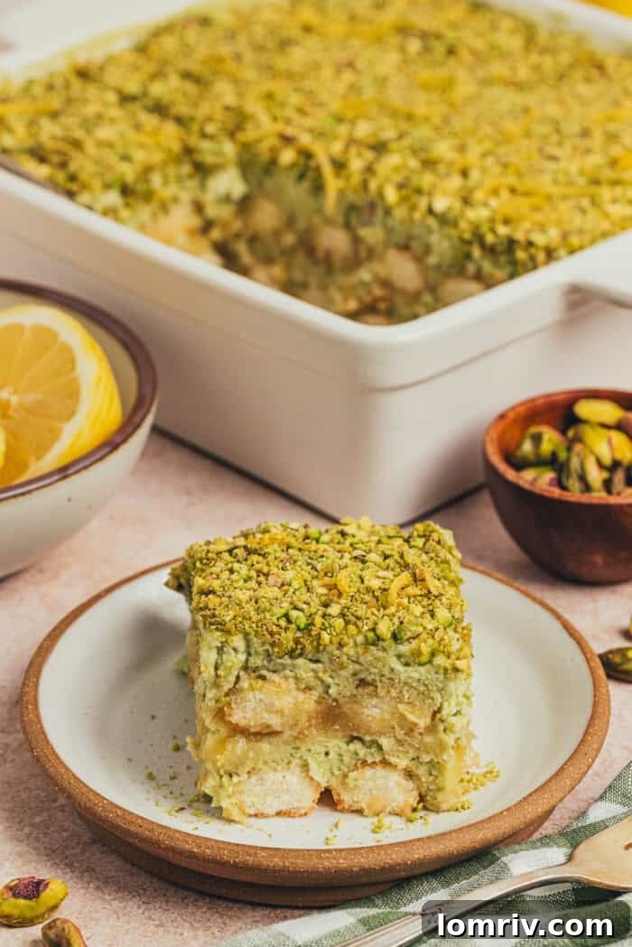 A slice of lemon pistachio tiramisu on a plate with a cake pan of tiramisu in the background.
